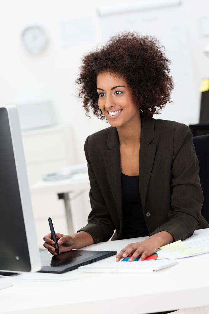 Creative young mixed race woman smiling while working as a designer at her desk, on a graphic tablet in front of a big monitor