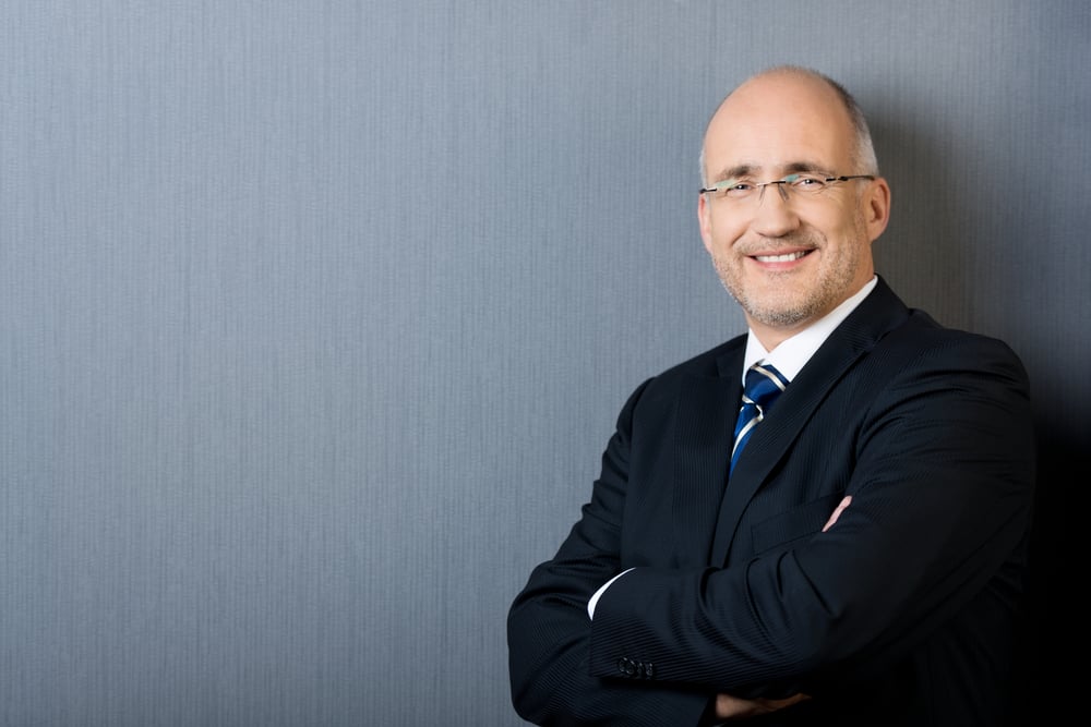 Profile portrait of a smiling and confident mature balding businessman, wearing a suit and a necktie, with arms crossed, in front of a gray wall with copy-space